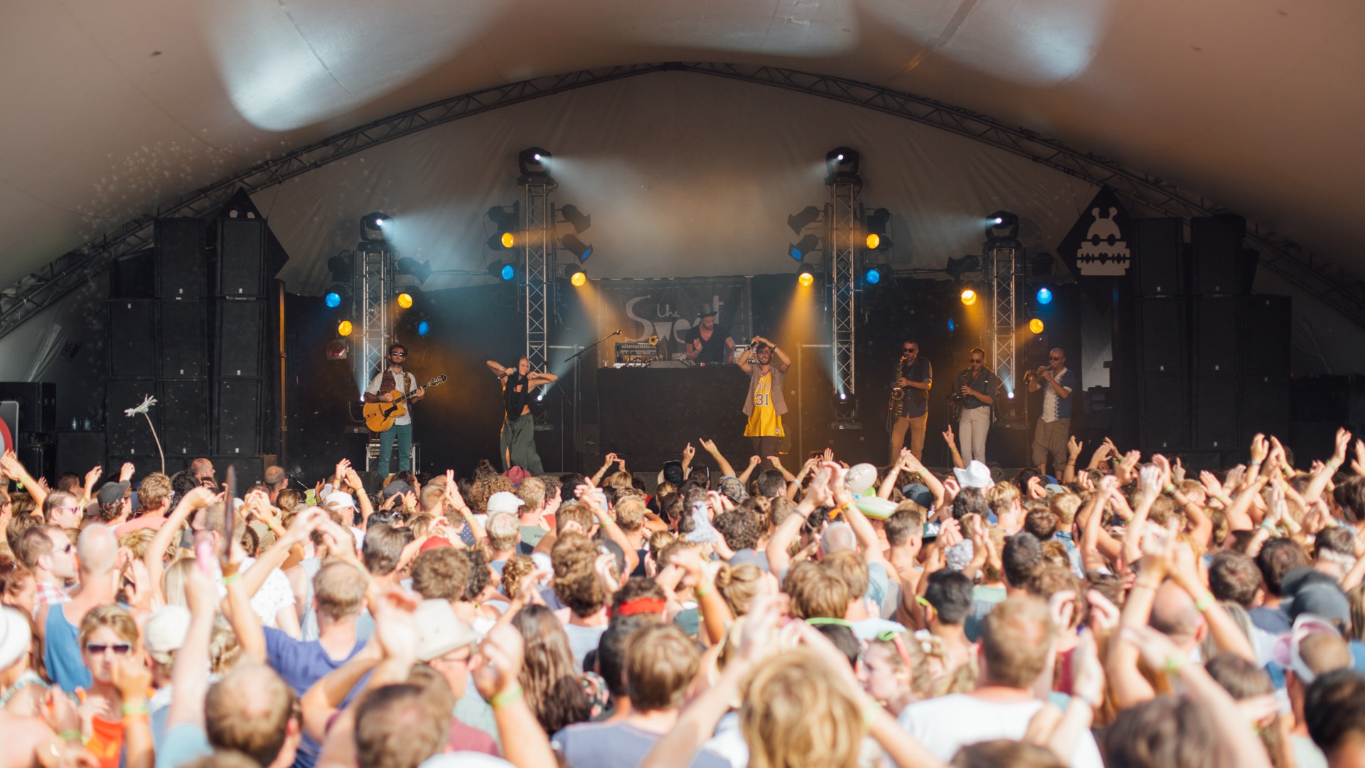 Daytime festival set under a stage canopy with the crowd's hands in the air